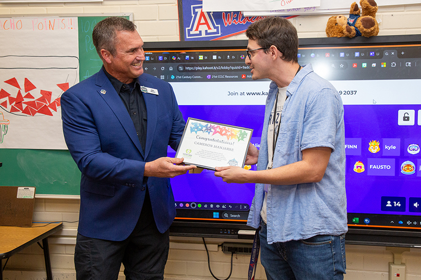 A man in a blue suit jacket smiles as he hands a certificate to a man in glasses and a blue button-down