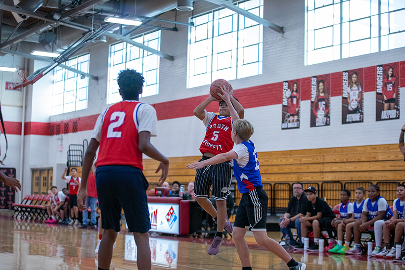 A boy in a red jersey jumps up to keep the basketball away from a boy in a blue jersey