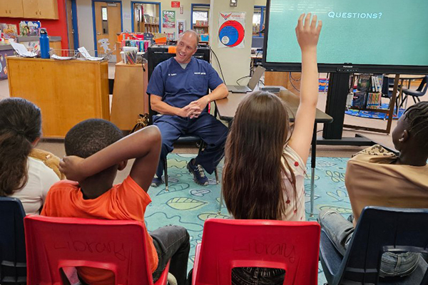 A man in blue scrubs sits next to a screen and answers students' questions