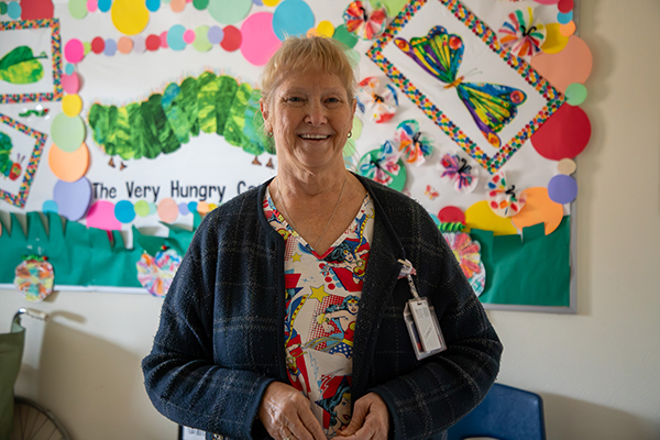 A woman in a plaid sweater and Wonder Woman shirt smiles in front of a board decorated with The Very Hungry Caterpillar