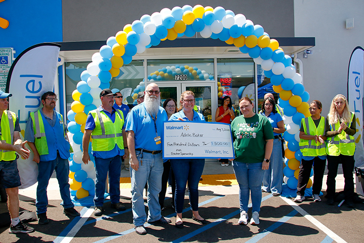 A woman in a blue shirt smiles as she holds a giant check next to a woman in a green shirt, under a blue, yellow and white balloon arch